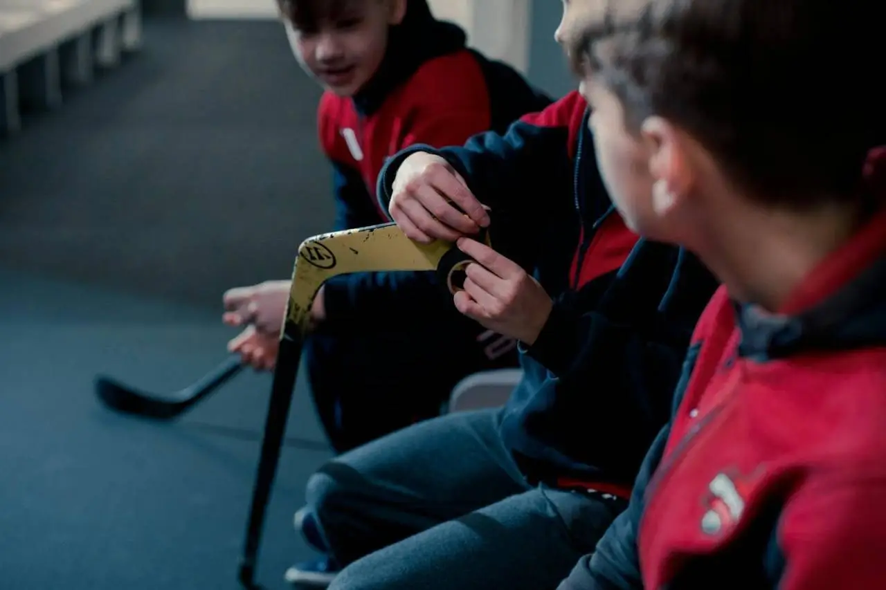 Children in a locker room taping hockey sticks, preparing for a game.