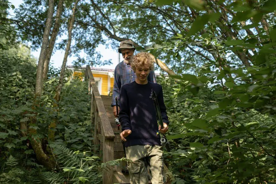 A grandfather and his grandson walk together in a lush green forest, carrying fishing gear.