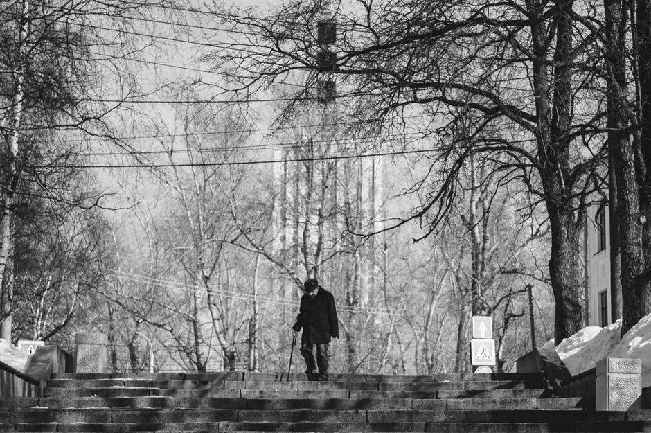 Monochrome photo of an elderly man with a cane walking down concrete stairs in Arkhangelsk city on a winter day.