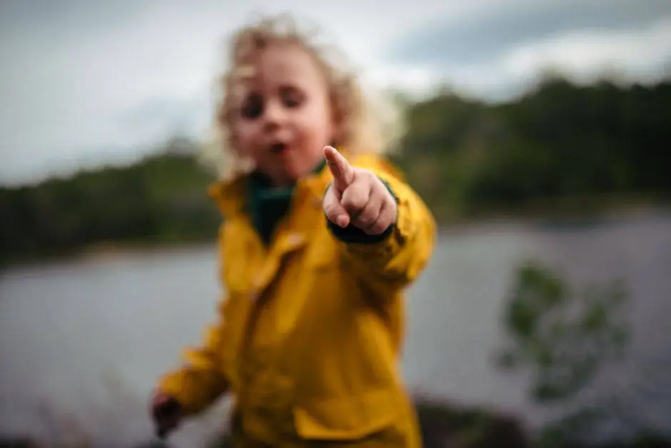 A young boy in a yellow jacket excitedly points while exploring the outdoors near a lake.